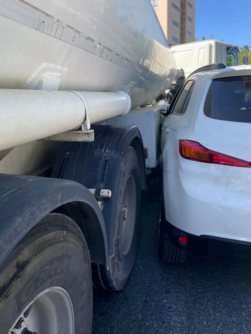 A white car is wedged between the wheels of a semi-truck after a likely blind spot accident. 